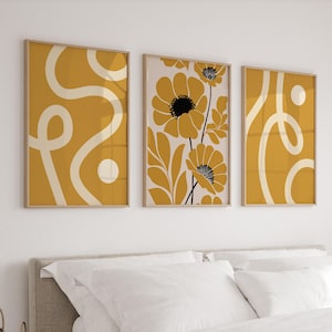 Può includere: Tre stampe artistiche incorniciate con un'estetica moderna della metà del secolo. Le stampe presentano disegni astratti in giallo senape e crema, e motivi floreali con accenti neri. Le cornici sono di colore legno chiaro, sopra un letto.