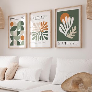 May include: Three framed art prints with abstract designs. The prints feature geometric shapes, botanical illustrations, and the name "MATISSE." Colors include green, orange, and beige. The frames are light brown, and the prints are displayed above a white couch.