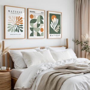 May include: Three framed art prints with abstract designs hang above a bed. The prints feature botanical and geometric shapes in orange, green, and white against neutral backgrounds. The text "MATISSE" and "BAUHAUS" are visible on the prints.