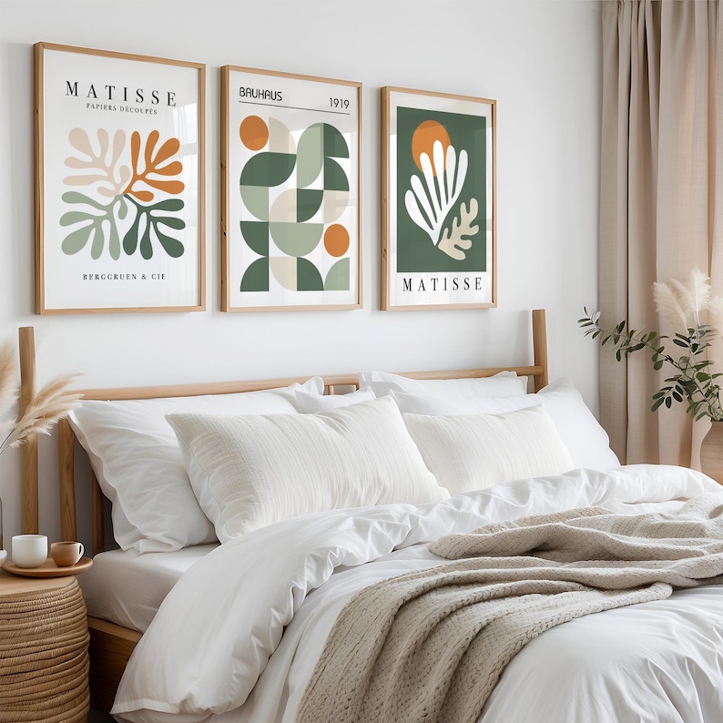 May include: Three framed art prints with abstract designs hang above a bed. The prints feature botanical and geometric shapes in orange, green, and white against neutral backgrounds. The text "MATISSE" and "BAUHAUS" are visible on the prints.