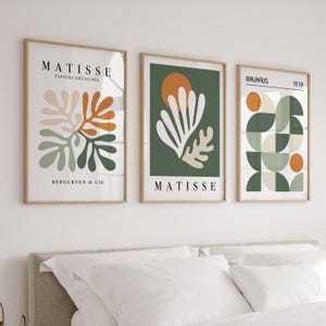 May include: Three framed art prints with abstract designs. The prints feature neutral colors, including beige, orange, green, and white, with the text "MATISSE" and "BAUHAUS" displayed. The art is displayed above a bed with white bedding.