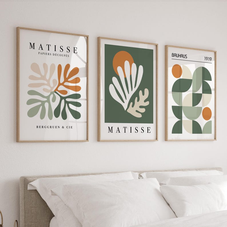 May include: Three framed art prints with abstract designs. The prints feature neutral colors, including beige, orange, green, and white, with the text "MATISSE" and "BAUHAUS" displayed. The art is displayed above a bed with white bedding.
