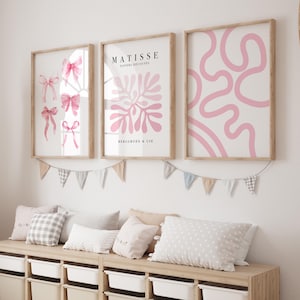 K&ouml;nnte beinhalten: Drei gerahmte Kunstdrucke mit rosafarbenen abstrakten Designs. Die Drucke h&auml;ngen an einer wei&szlig;en Wand mit einer Girlande aus pastellfarbenen Flaggen darunter.