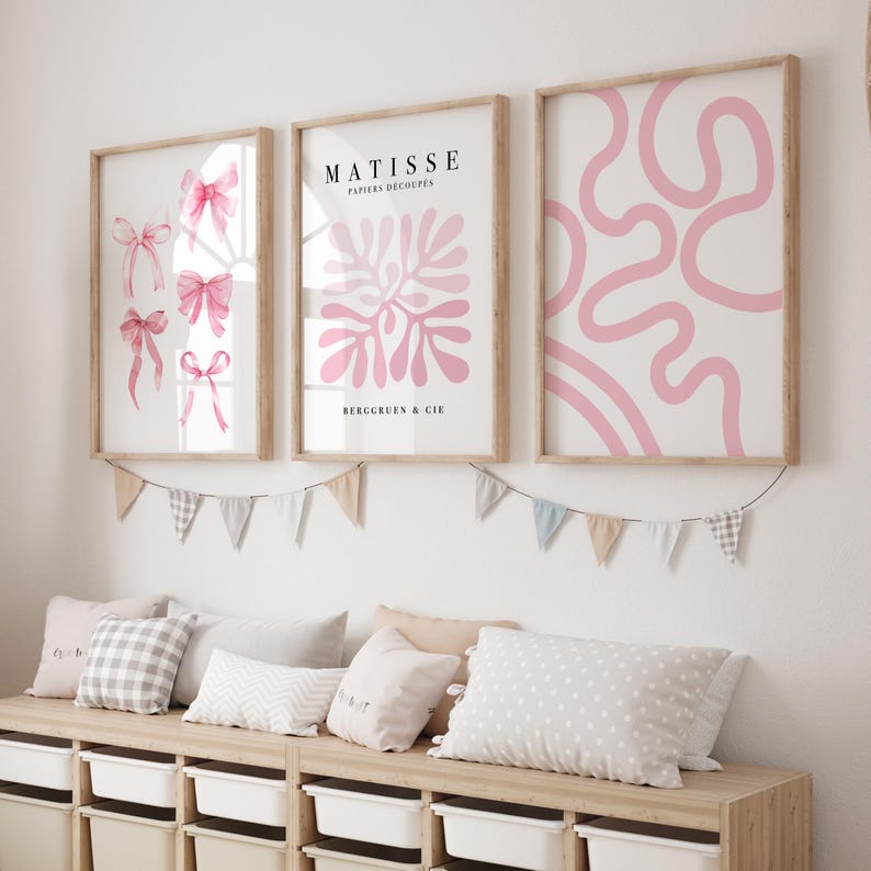 K&ouml;nnte beinhalten: Drei gerahmte Kunstdrucke mit rosafarbenen abstrakten Designs. Die Drucke h&auml;ngen an einer wei&szlig;en Wand mit einer Girlande aus pastellfarbenen Flaggen darunter.