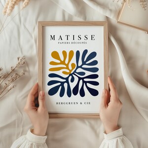 Puede incluir: Una impresi&oacute;n enmarcada con un dise&ntilde;o abstracto inspirado en Matisse, que presenta formas azules y amarillas. La impresi&oacute;n dice "Matisse Papiers D&eacute;coup&eacute;s" y "Berggruen & Cie".