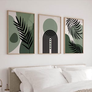 Peut inclure: Trois impressions d'art abstraites encadrées. Chaque impression est encadrée dans du bois clair. L'art comprend des formes vertes et blanches, des silhouettes de feuilles noires et un motif en arc noir et blanc. Les impressions sont affichées au-dessus d'un lit avec une literie blanche.
