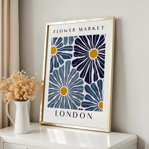 Puede incluir: Una impresi&oacute;n enmarcada con un patr&oacute;n floral azul y blanco y centros amarillos. La impresi&oacute;n dice "FLOWER MARKET LONDON" en la parte superior e inferior.