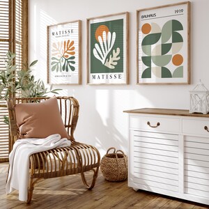 May include: Three framed art prints with abstract designs hang on a white wall. The prints feature botanical and geometric shapes in shades of green, orange, and white. The word "MATISSE" is printed on two of the prints. A wicker chair, pillow, and blanket are in the foreground.