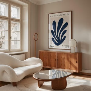 Puede incluir: Un sof&aacute; blanco curvo se encuentra en una sala de estar con un gabinete de madera y una mesa de caf&eacute; de vidrio. Una impresi&oacute;n enmarcada de un dise&ntilde;o abstracto azul con el texto "PAPIERS DECOUPES H. MATISSE" cuelga de la pared.
