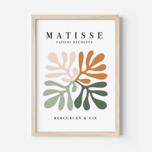 May include: A framed art print featuring a colorful abstract design. The print displays leaf-like shapes in shades of beige, orange, and green, arranged in a square. The text "MATISSE PAPIERS D&Eacute;COUP&Eacute;S" and "BERGGRUEN & CIE" are printed in black.
