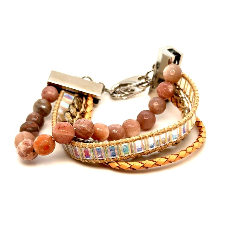 Op de afbeelding: Een armband met meerdere strengen met zonnesteen kralen en een gevlochten leren band. De armband heeft een zilverkleurige sluiting en een band met iriserende rechthoekige kralen. De zonnesteen kralen zijn een mix van oranje en bruin.
