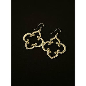 May include: A pair of silver-coloured, openwork earrings. Each earring features a quatrefoil design with a hook for pierced ears. The earrings have a decorative, ornate style, with a smooth, polished finish. The background is black.
