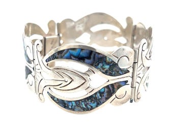 May include: A silver bracelet with a hinged design. The bracelet features a repeating pattern of stylized leaves and oval inlays of iridescent blue and green abalone shell. The bracelet is shown against a white background.
