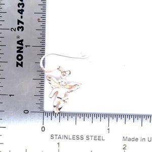 May include: A silver-colored angel-shaped earring with detailed wings and a hook. The earring is shown next to a ruler with measurements in inches. The text "STAINLESS STEEL" is visible.