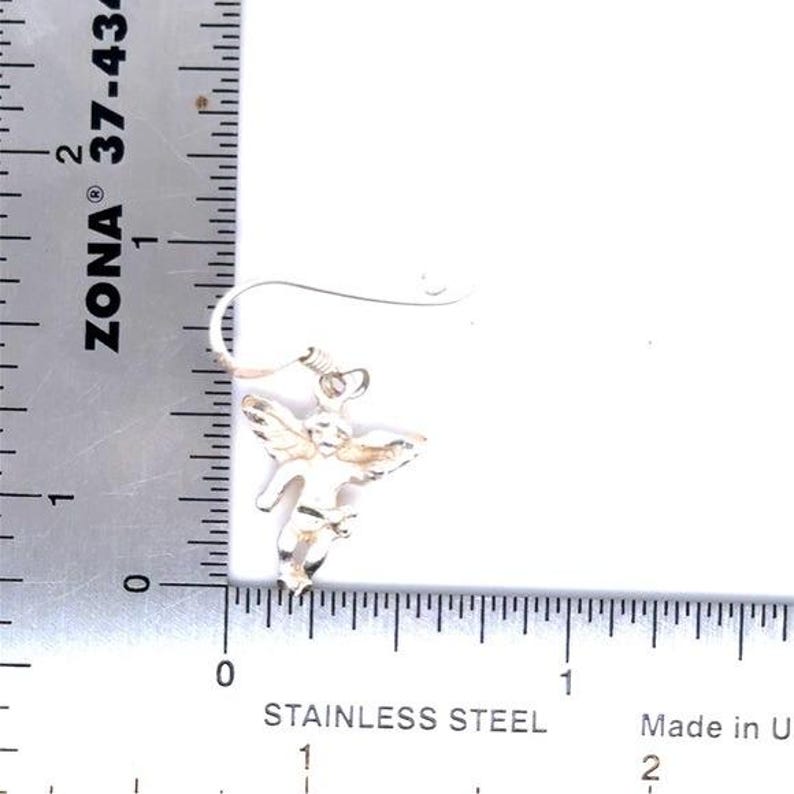 May include: A silver-colored angel-shaped earring with detailed wings and a hook. The earring is shown next to a ruler with measurements in inches. The text "STAINLESS STEEL" is visible.