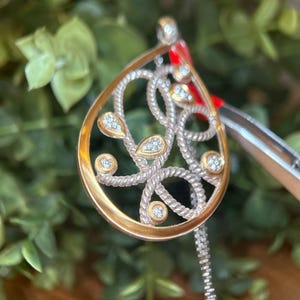 May include: A gold and silver pendant in a teardrop design. The pendant has a gold edge, silver rope-style detailing, and multiple small, round, sparkling gemstones. A silver chain is attached to the pendant.