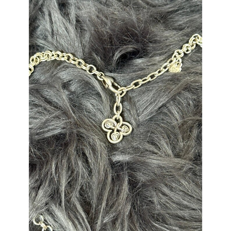 May include: Silver-toned necklace with a clover-shaped pendant. The pendant has four heart-shaped designs, each with a small, clear stone. The chain is made of small, oval links and has a clasp closure.