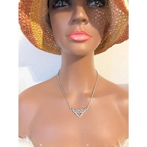 May include: A silver-toned necklace with a Celtic knot pendant. The pendant is a heart-shaped design with interwoven lines. The necklace is displayed on a mannequin wearing a straw hat with orange and yellow tones.