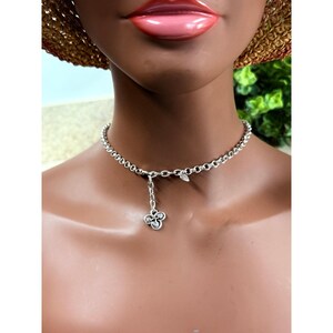 May include: A silver-toned choker necklace with a delicate chain design. The necklace features a small pendant with a clover-like design. The necklace is worn around the neck.