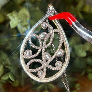 May include: Silver pendant necklace with a teardrop design. The pendant has an openwork pattern with a rope-like texture and small, round details. The necklace is shown against a blurred green background, with a silver chain.