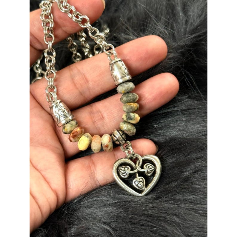 May include: A silver-toned necklace featuring a chain and a pendant. The necklace has a heart-shaped pendant with leaf designs and a strand of small, colorful stones. The chain is made of interlocking links.
