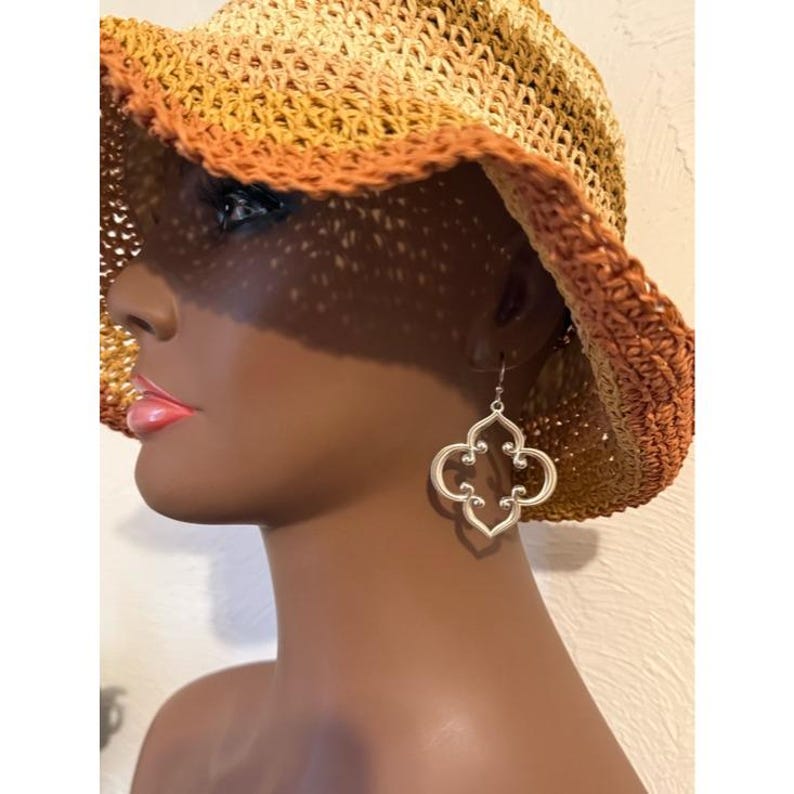 May include: A silver-coloured, openwork quatrefoil earring hangs from a mannequin's ear. The earring is a stylised floral design. The mannequin is wearing a straw hat with a brown and orange gradient.