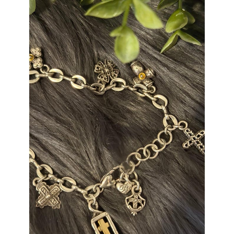 May include: Silver-toned charm bracelet with various religious-themed charms. The bracelet features a chain link design with multiple pendants, including crosses, floral designs, and a rectangular charm with gold accents. The charms are detailed and have a vintage aesthetic.