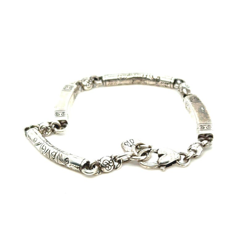 Puede incluir: Una pulsera de plata con un dise&ntilde;o ornamentado. La pulsera presenta una serie de eslabones rectangulares y cil&iacute;ndricos, cada uno adornado con intrincados patrones. Un cierre en forma de coraz&oacute;n asegura la pulsera. La joyer&iacute;a se muestra sobre un fondo blanco.