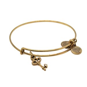May include: A gold-toned bangle bracelet with a key charm and two smaller charms. The key charm has a small crystal accent. The bracelet is made of metal and has a simple, elegant design. The charms include a clover and a charm with an American flag.