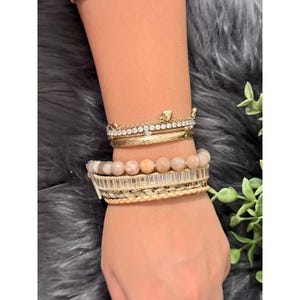 Op de afbeelding: Een verzameling gouden armbanden en een kralenarmband zijn op een pols gestapeld. De gouden armbanden omvatten een kroonvormige armband met heldere stenen en een getextureerde band. De kralenarmband heeft lichtroze stenen en heldere rechthoekige kralen.