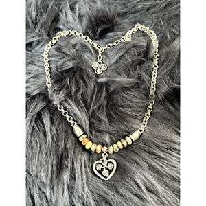 May include: Silver-toned necklace with a heart pendant and a beaded section. The heart pendant has a leaf design inside. The necklace is on a gray, furry surface.