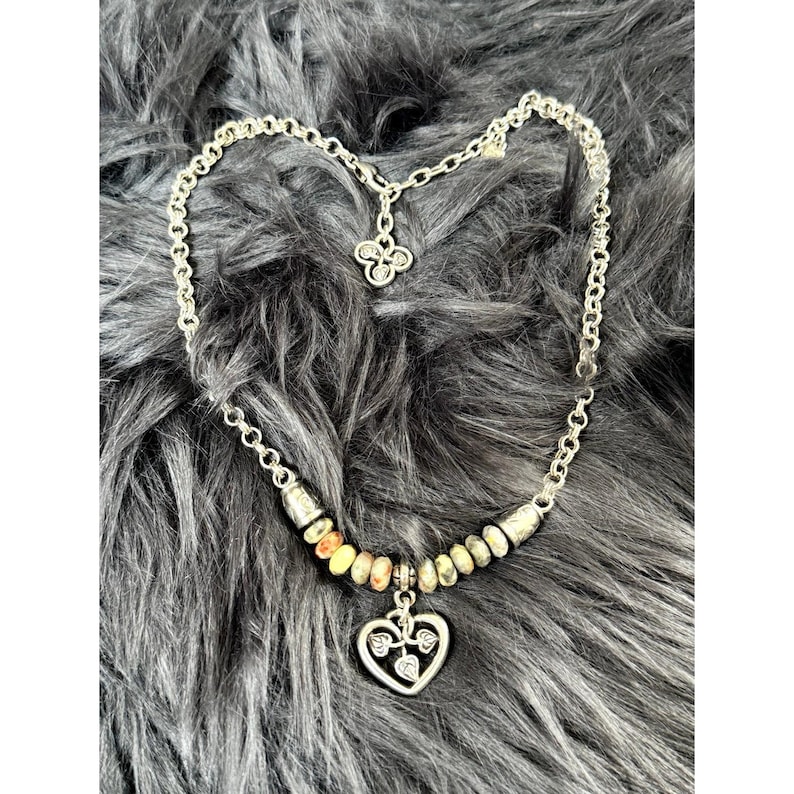 May include: Silver-toned necklace with a heart pendant and a beaded section. The heart pendant has a leaf design inside. The necklace is on a gray, furry surface.
