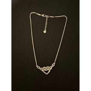 May include: A silver-toned necklace with a heart-shaped pendant. The pendant is formed by intertwined loops, creating a knot design. The necklace has a delicate chain and an adjustable clasp.
