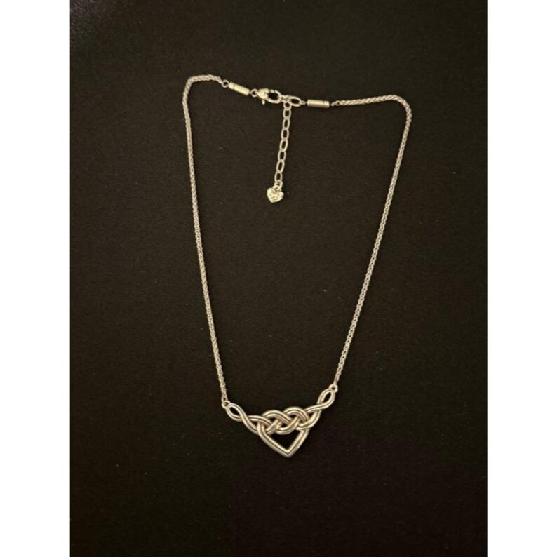 May include: A silver-toned necklace with a heart-shaped pendant. The pendant is formed by intertwined loops, creating a knot design. The necklace has a delicate chain and an adjustable clasp.