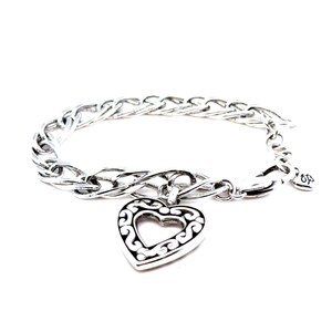 Op de afbeelding: Zilverkleurige armband met een hartvormige hanger met een open ontwerp. De hanger hangt aan een schakelarmband. De armband heeft een karabijnsluiting en een kleine decoratieve bedel.