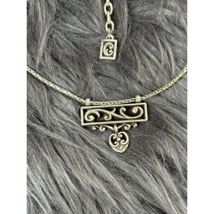 May include: A silver-toned necklace with a rectangular pendant featuring an openwork design of swirling patterns and a heart-shaped charm. The necklace has a delicate chain and a matching adjustable extension.