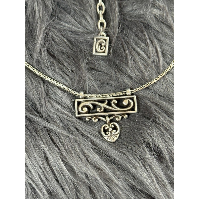 May include: A silver-toned necklace with a rectangular pendant featuring an openwork design of swirling patterns and a heart-shaped charm. The necklace has a delicate chain and a matching adjustable extension.