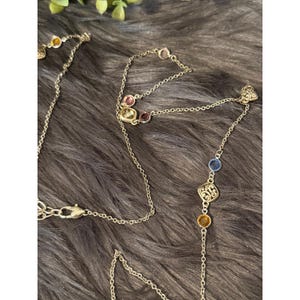 K&ouml;nnte beinhalten: Eine silberfarbene Halskette mit farbigen Edelsteinen. Die Halskette hat eine filigrane Kette mit verschiedenfarbigen Steinen, darunter Gelb, Rosa und Blau. Die Edelsteine sind in eine dekorative Fassung gesetzt.