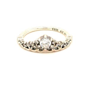 May include: A silver-colored ring with a crown-like design. The ring features a large, clear, round gemstone at the center, surrounded by smaller, clear gemstones. The band is thin and has the text "S925 ALE" engraved on it.