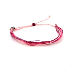 May include: A multi-strand string bracelet featuring various shades of pink, magenta, and purple threads. The bracelet includes a small dark charm with a white logo and an adjustable sliding knot closure with two loose ends extending from the center.