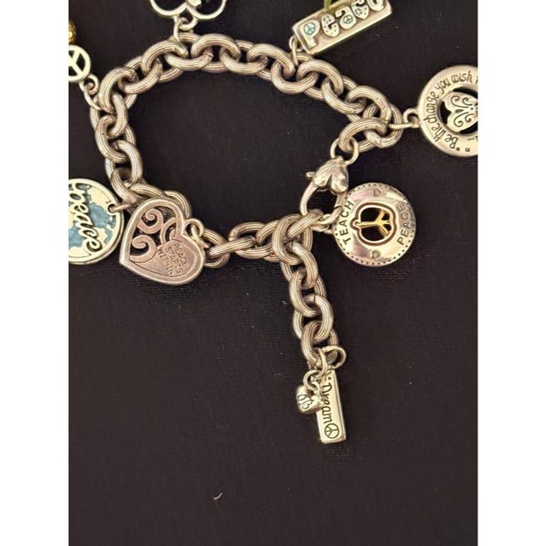 May include: Silver-toned charm bracelet with multiple charms. Charms include a heart with a filigree design, a round charm with a peace symbol, and a rectangular charm with the word "peace". Other charms have the words "Dream" and "Be the change you wish".
