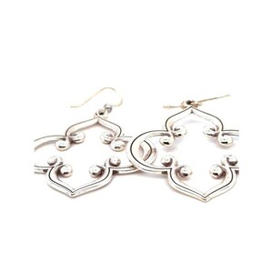 May include: A pair of silver earrings with an openwork quatrefoil design. Each earring has small, round accents and a hook closure. The earrings are displayed against a white background. The design is symmetrical.