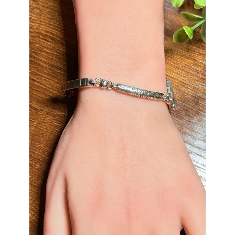 Puede incluir: Una pulsera plateada con una barra rectangular y un patr&oacute;n floral decorativo. La pulsera est&aacute; en una mu&ntilde;eca y el cierre es de estilo barra en T. La pulsera est&aacute; hecha de un metal brillante.