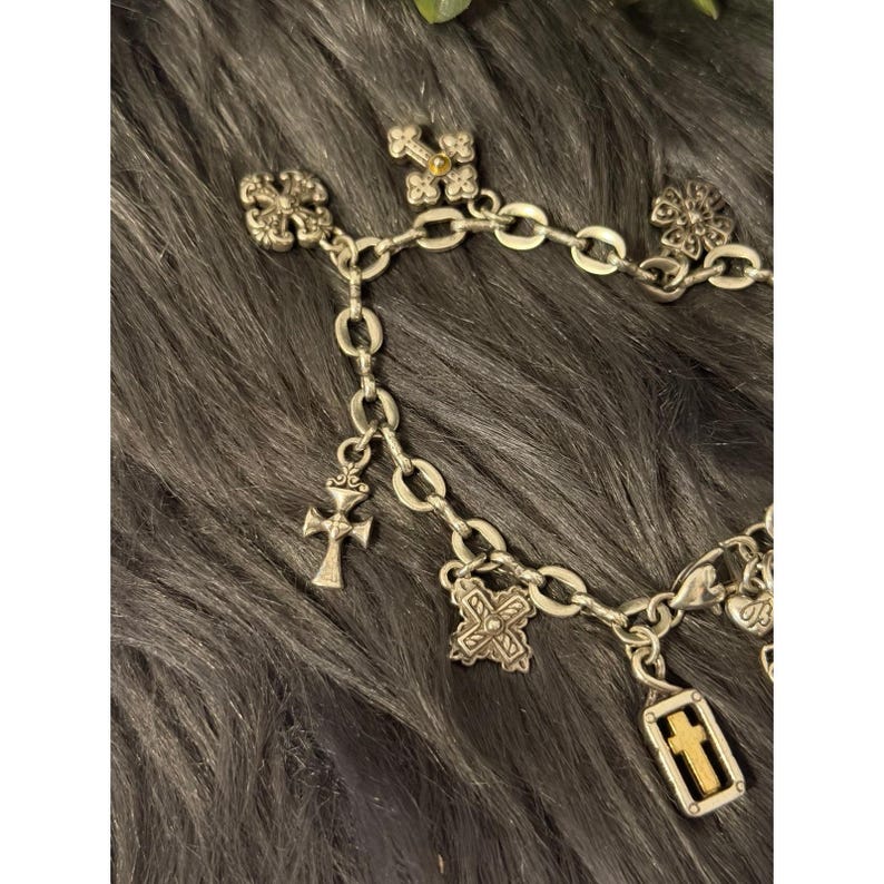 May include: Silver-toned charm bracelet featuring various cross and floral charms. The bracelet has a chain link design with multiple decorative elements, including crosses and ornate floral accents.