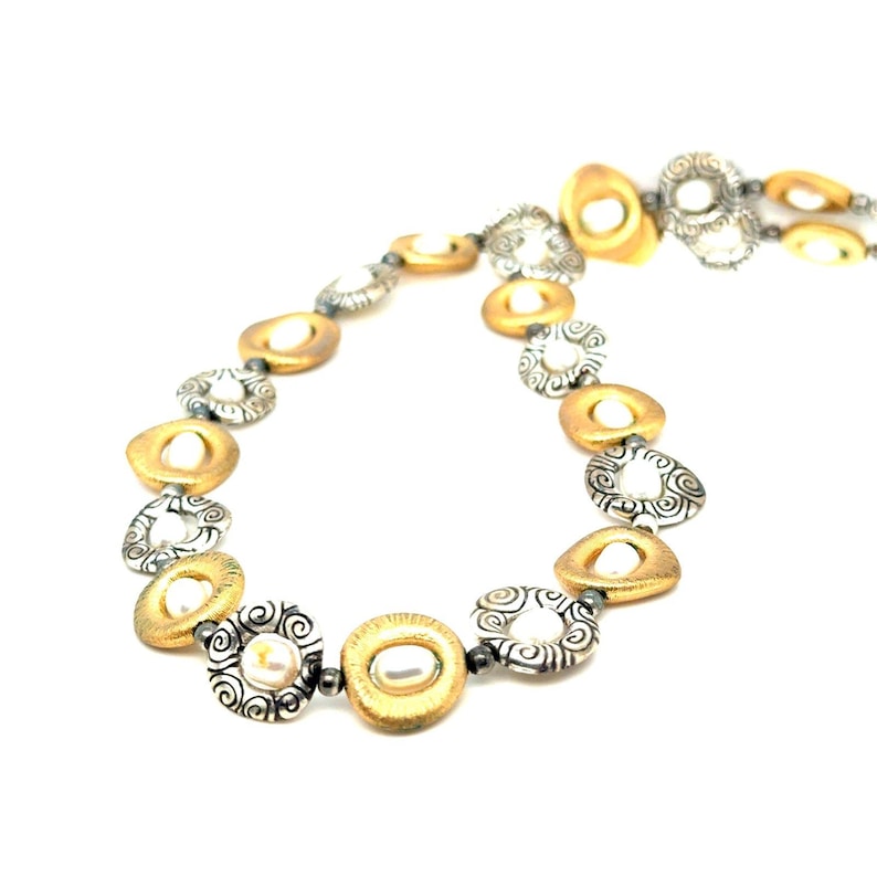 Puede incluir: Un collar dorado y plateado con un patr&oacute;n repetitivo de formas circulares y ovaladas. El collar presenta colgantes dorados de forma ovalada con un centro similar a una perla, intercalados con colgantes circulares calados plateados.