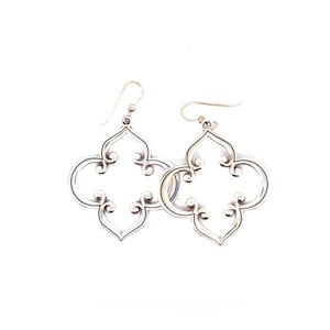 May include: A pair of silver-coloured, openwork earrings. Each earring features a quatrefoil design with curved edges and decorative swirls. The earrings have a hook closure and are displayed against a white background.
