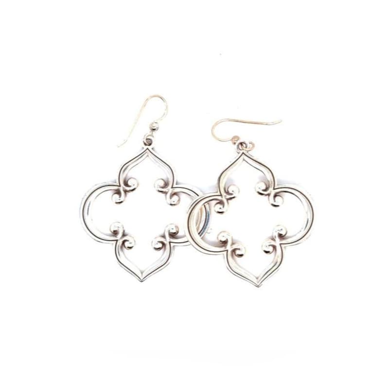May include: A pair of silver-coloured, openwork earrings. Each earring features a quatrefoil design with curved edges and decorative swirls. The earrings have a hook closure and are displayed against a white background.