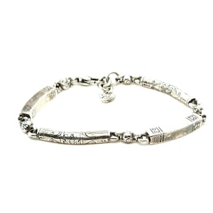 Puede incluir: Pulsera de plata con detalles decorativos grabados. La pulsera presenta eslabones rectangulares y cil&iacute;ndricos, conectados por eslabones m&aacute;s peque&ntilde;os con cuentas. Un dije en forma de coraz&oacute;n cuelga cerca del cierre. Sobre fondo blanco.