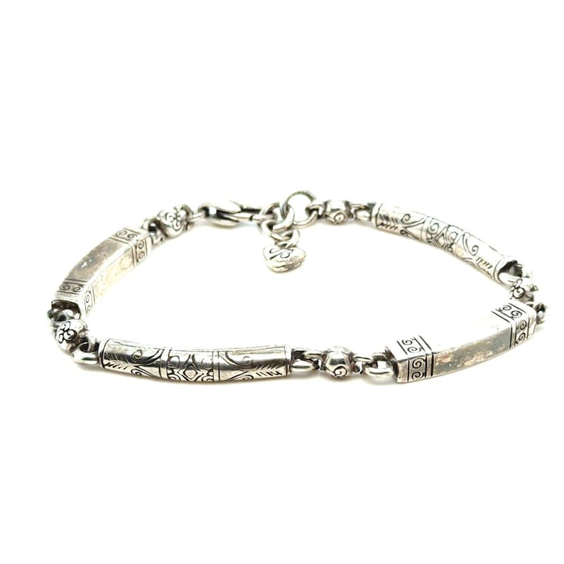 Puede incluir: Pulsera de plata con detalles decorativos grabados. La pulsera presenta eslabones rectangulares y cil&iacute;ndricos, conectados por eslabones m&aacute;s peque&ntilde;os con cuentas. Un dije en forma de coraz&oacute;n cuelga cerca del cierre. Sobre fondo blanco.