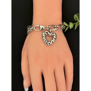 Op de afbeelding: Zilverkleurige armband met een hartvormige bedel. De bedel heeft een open ontwerp met ingewikkelde krullen. De armband wordt getoond op een mannequinhand, die de stijl van de sieraden benadrukt. De ketting is een curb link stijl.